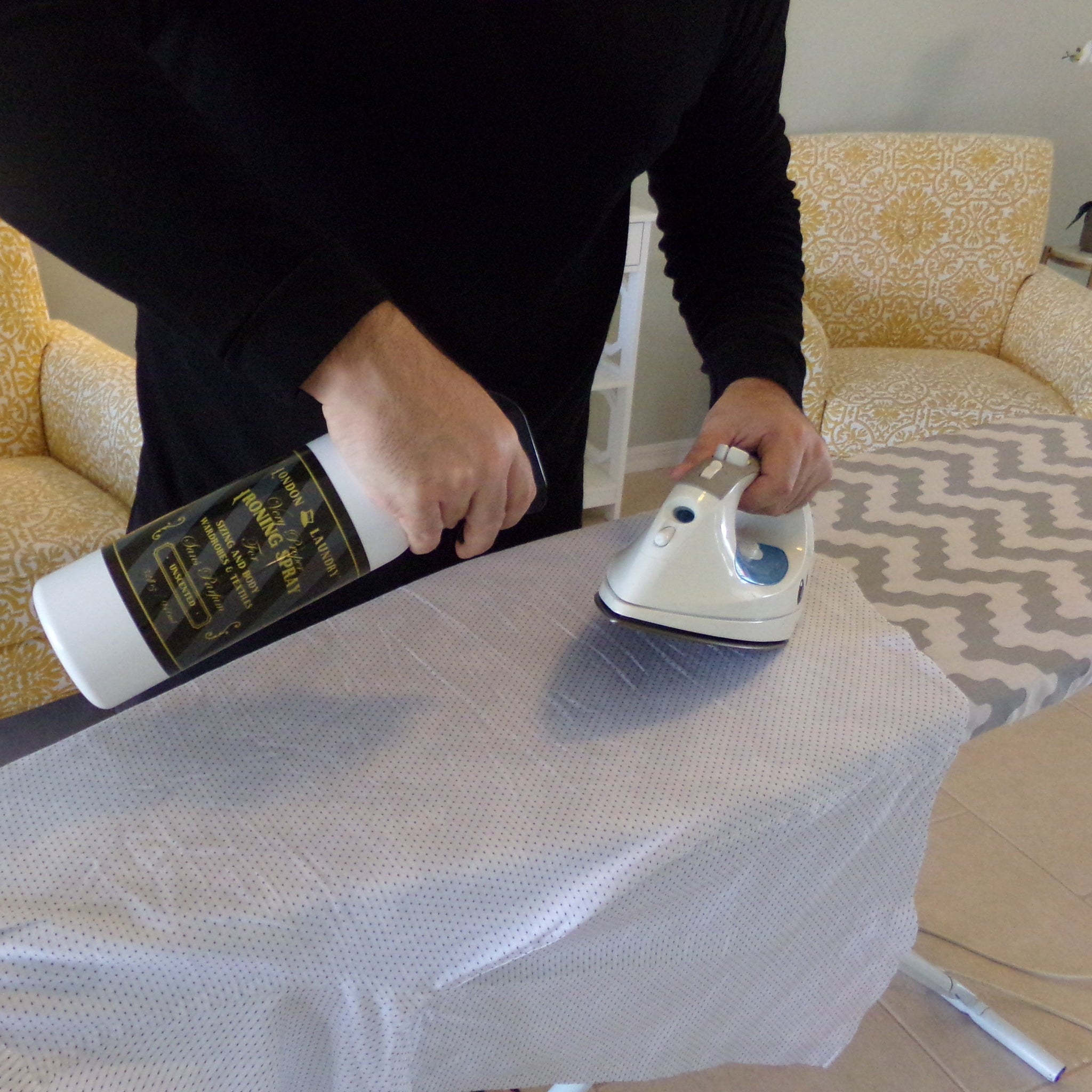 Person using a bottle and iron on an ironing board with yellow chairs in the background